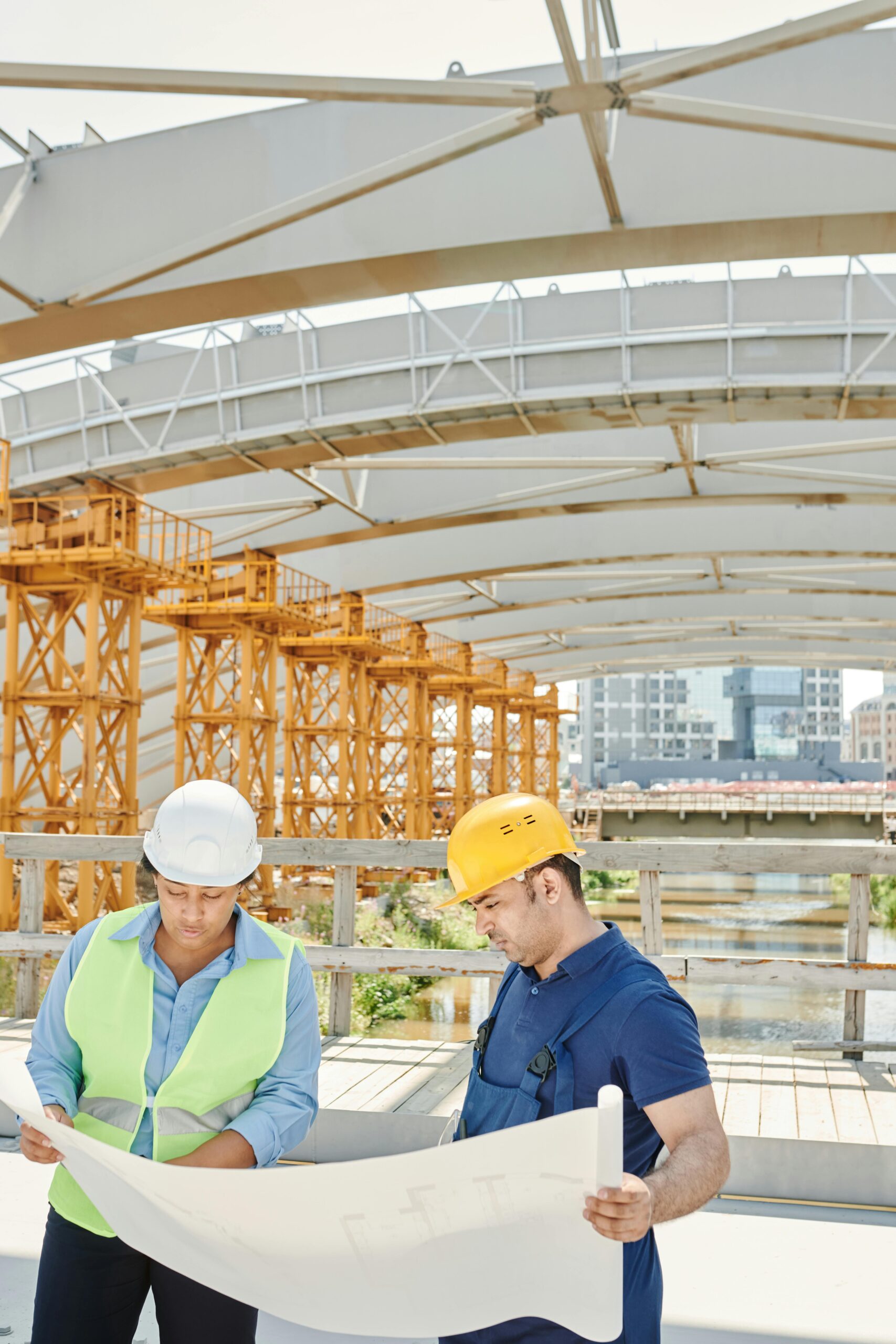 Two engineers on a construction site examining blueprints with steel structures in the background.